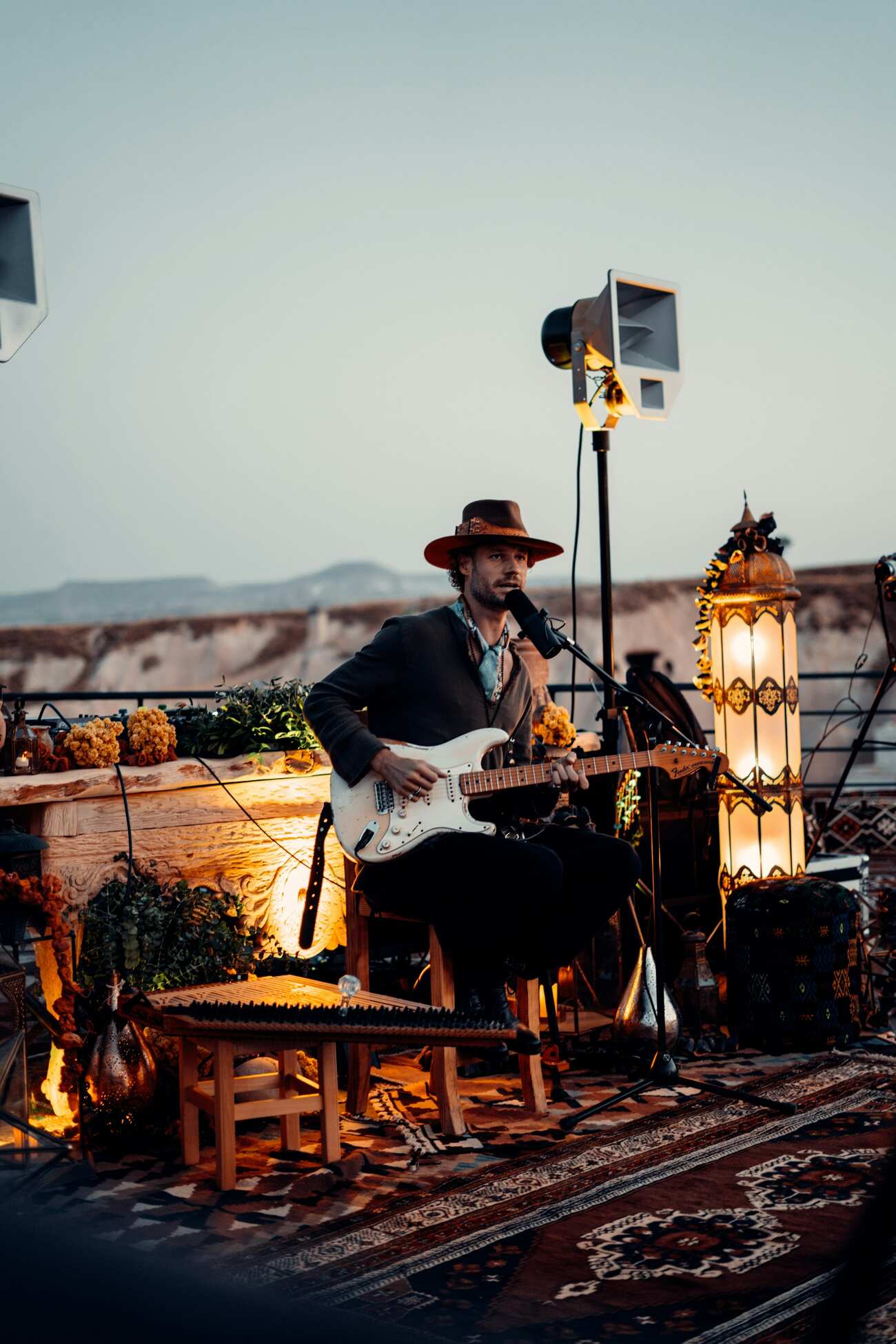 A man wearing a hat plays guitar on a rooftop, with a city skyline visible in the background