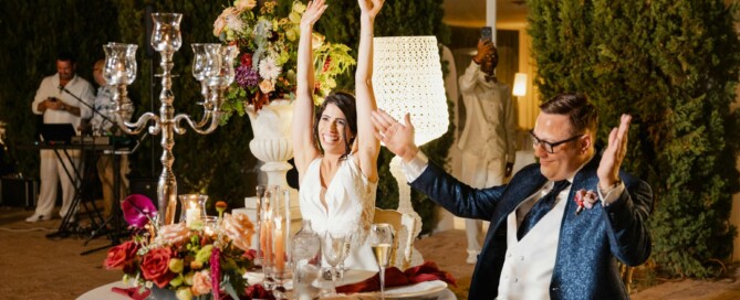 Bride and groom at sweetheart table raising their arms in celebration during reception