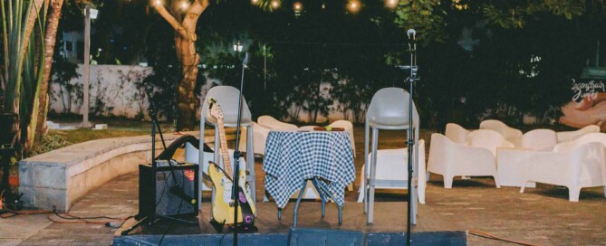 Empty outdoor performance setup with guitar, microphones, and chairs under hanging lights